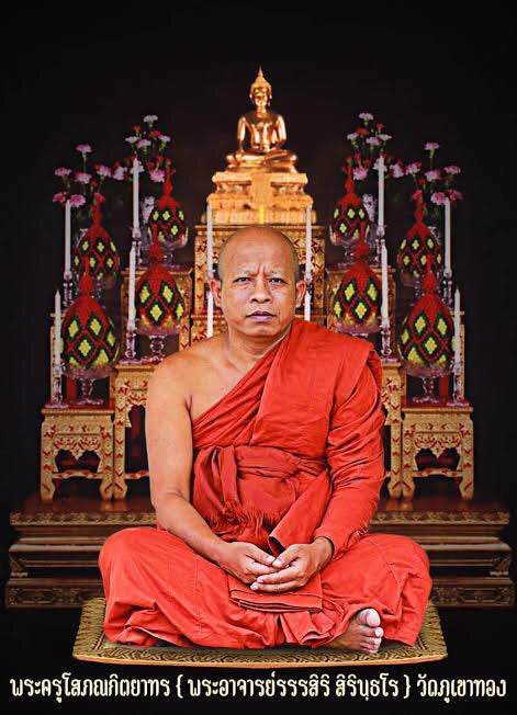 Man in orange robes seated before ornate shrine with Buddha statue and ceremonial ornaments presenting Great Axe Amulet Maha Prap
