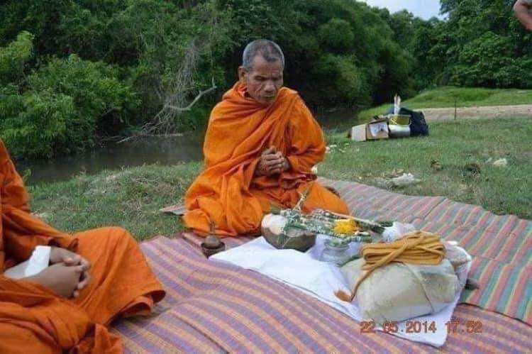 Hoon Payon ancient Thai guardian doll being blessed by monks outdoors on mat with offerings