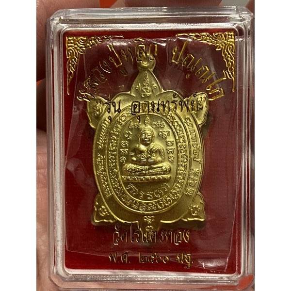 Gold Luang Pu Liew turtle amulet in plastic display case on red background showing seated monk and Thai script