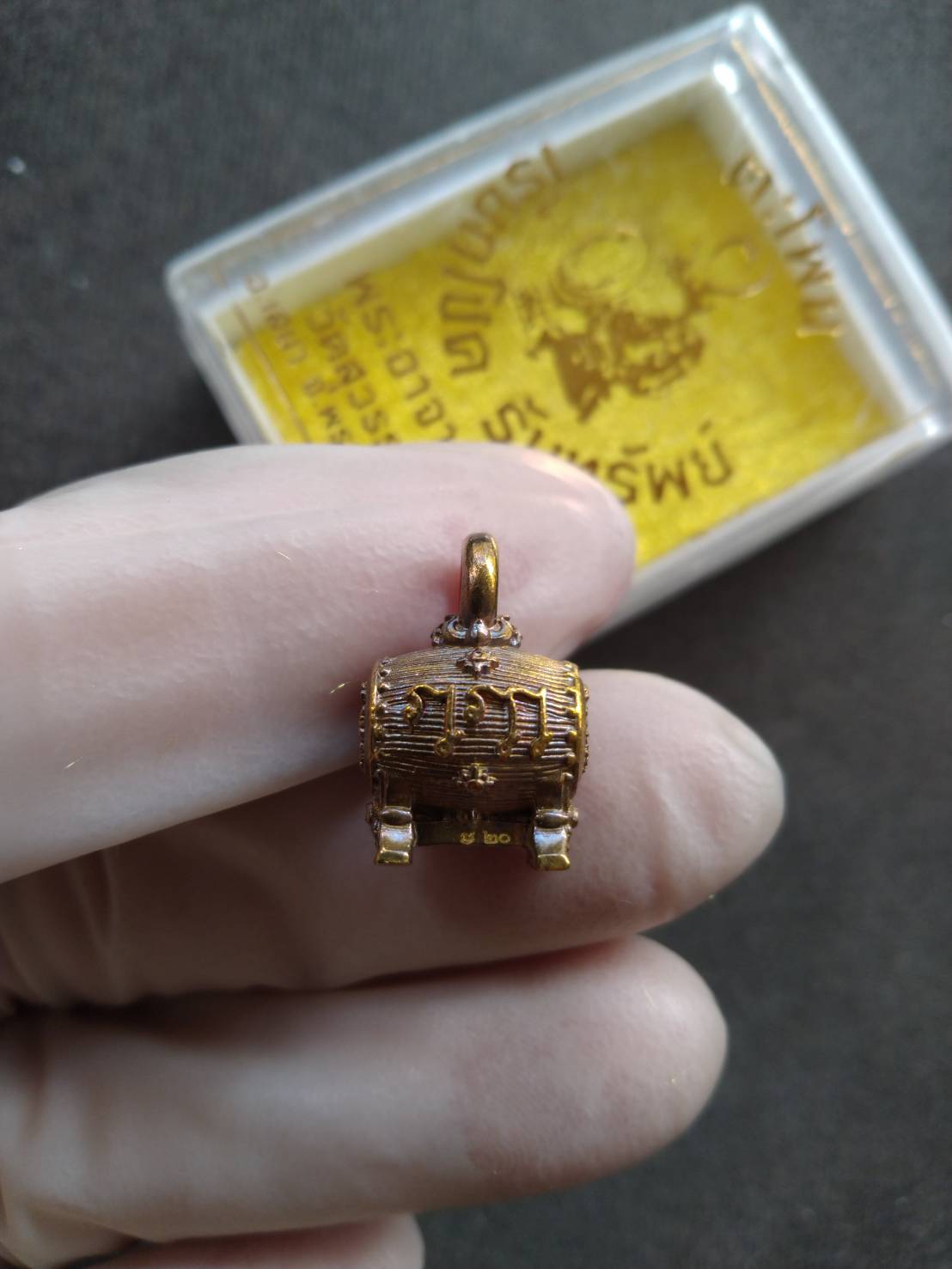 Thai Blessing Co gold tone Thai Buddha pendant held in gloved fingers with case in background Lucky amulet