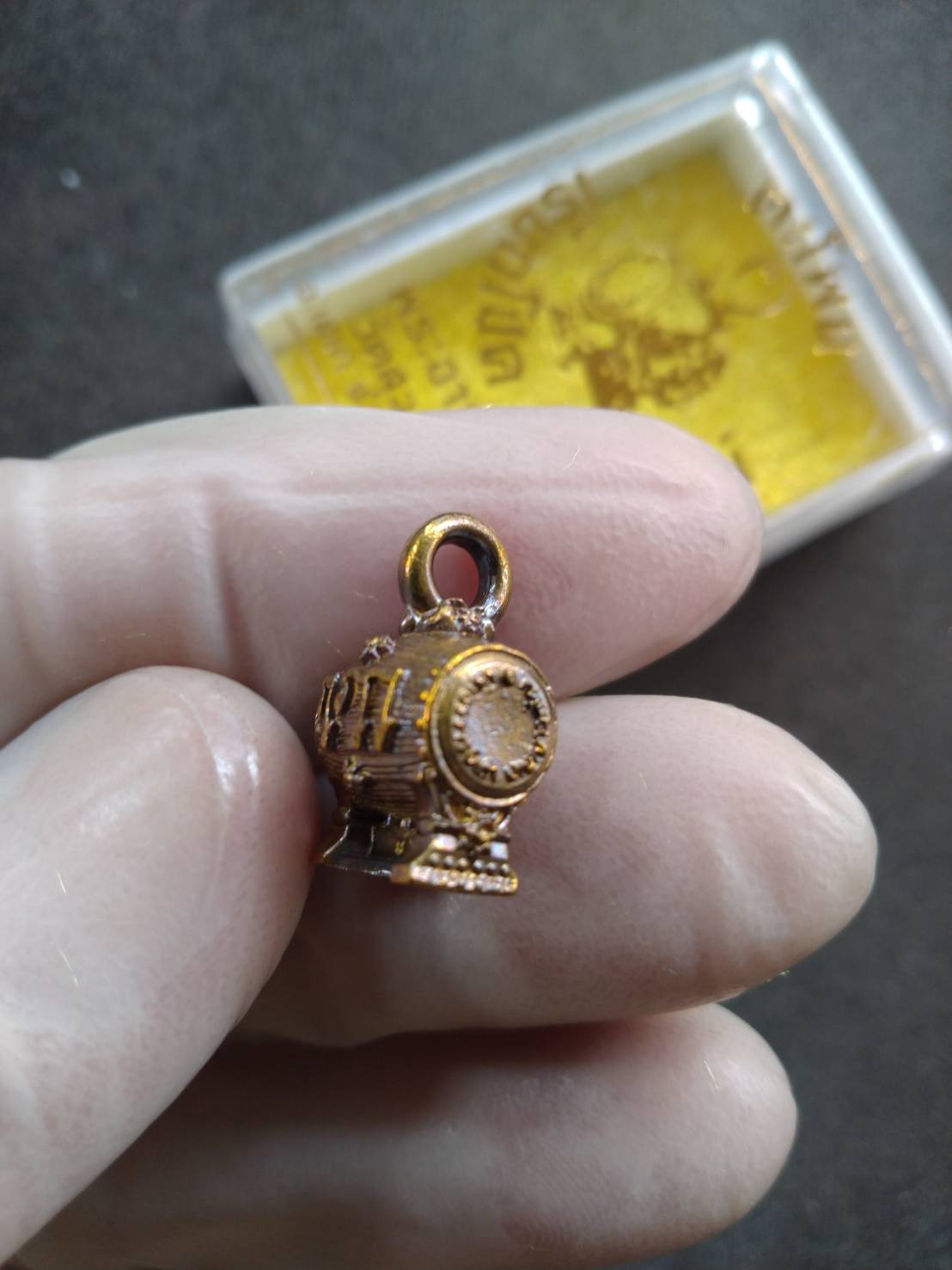 Thai Blessing Co gold miniature Buddhist pendant held in gloved fingers Lucky amulet with detailed carved face and loop bail