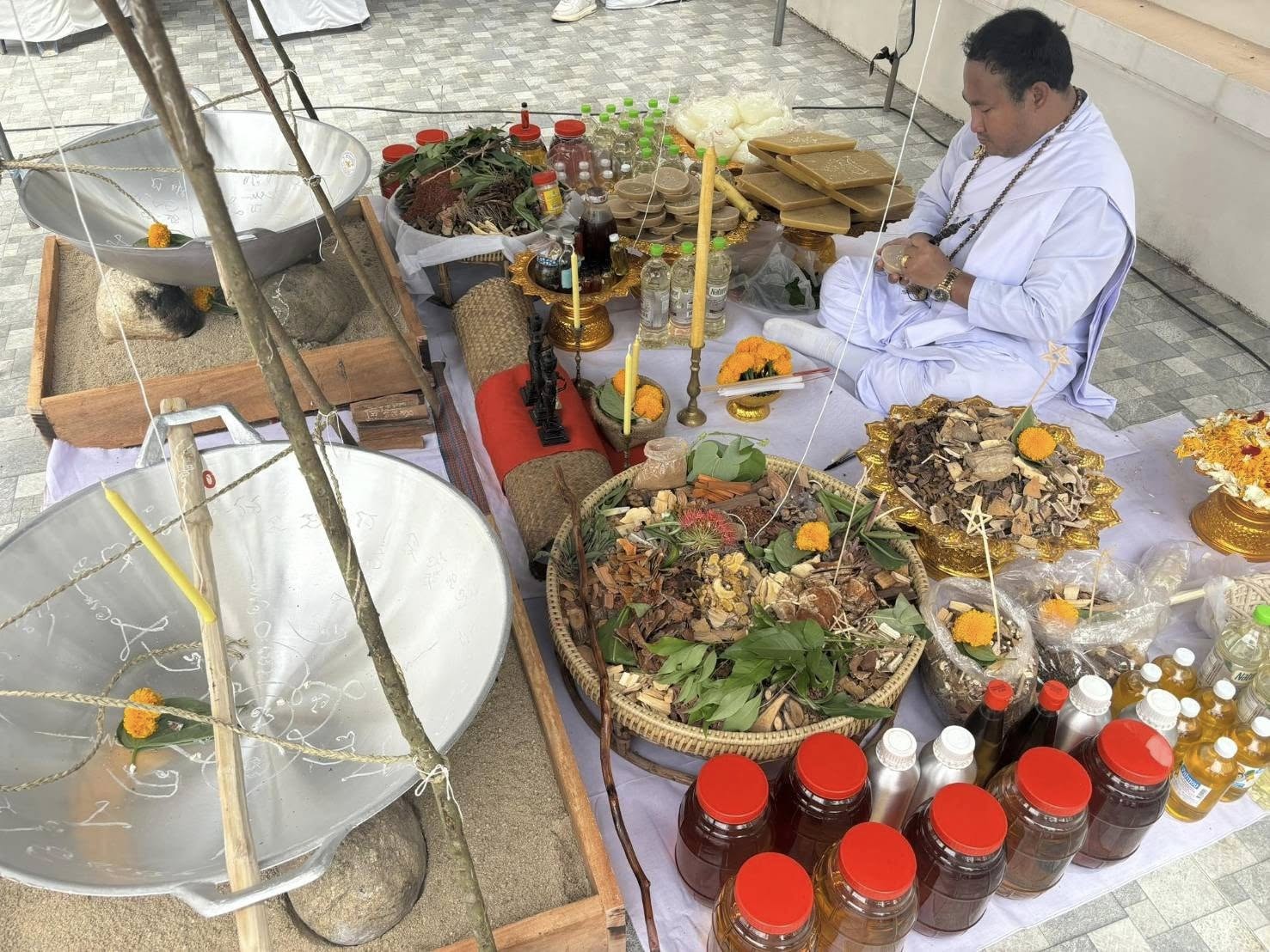 Thai Blessing Co priest in white robes performing Luuk Sawat sacred charm blessing with herbs jars marigolds