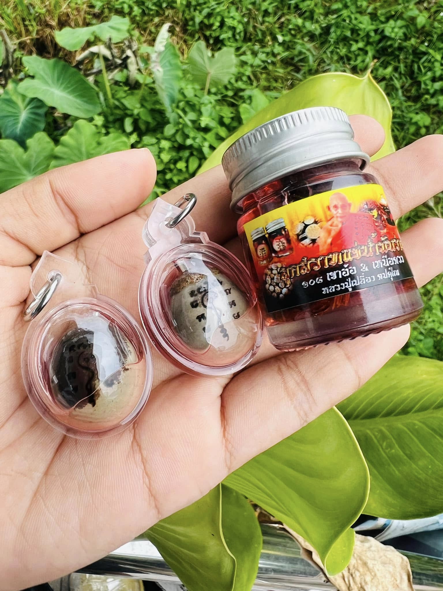 Hand holding two encased Luuk Sawat sacred charm amulets in plastic cases and a small Thai Blessing Co jar of red oil