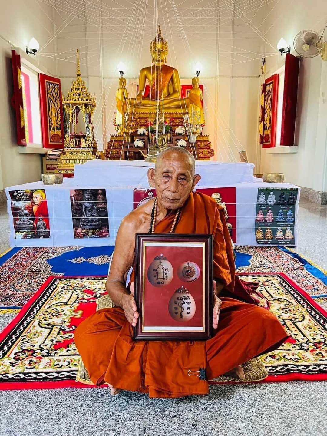 Thai Blessing Co monk holding framed Luuk Sawat sacred charm amulets seated in temple before golden Buddha
