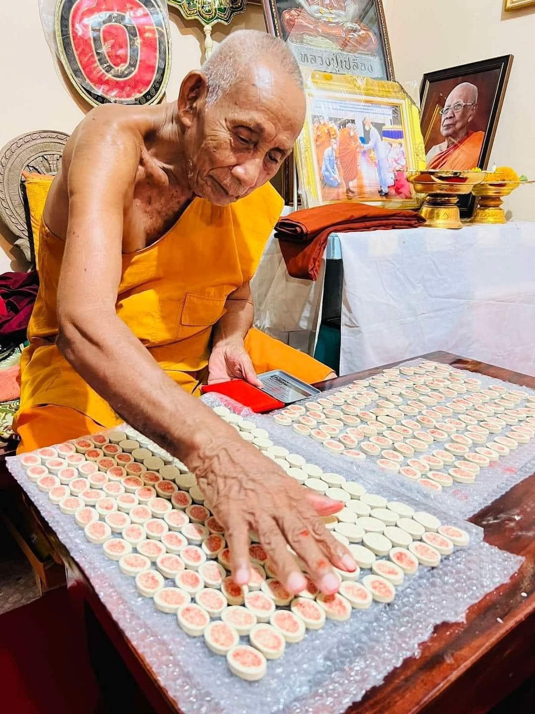Elderly Thai monk blessing rows of small round amulets on table Luuk Sawat sacred charm from Thai Blessing Co