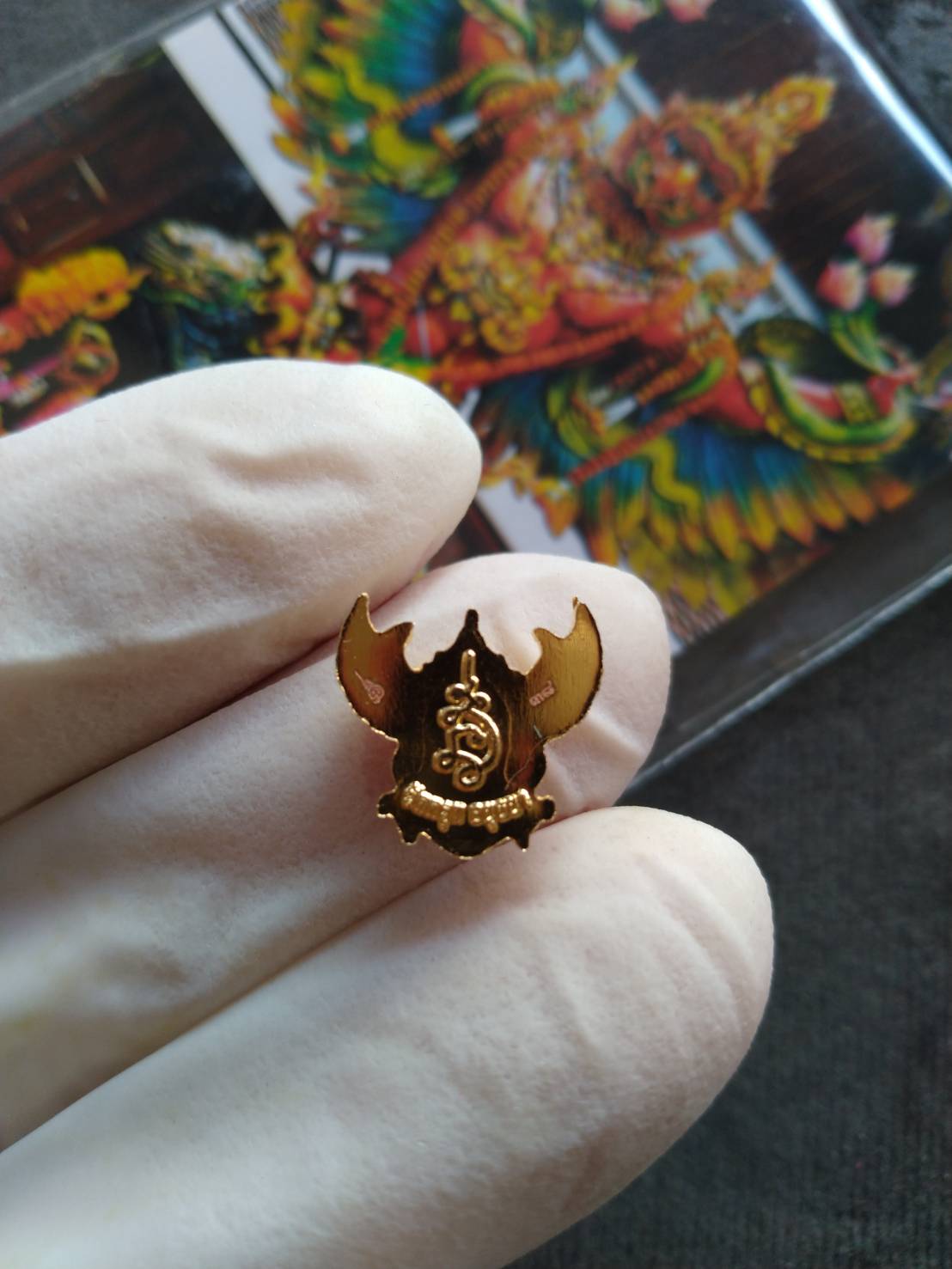 Close up of gold mini Garuda amulet pin pendant held by gloved fingers with colorful Garuda image background