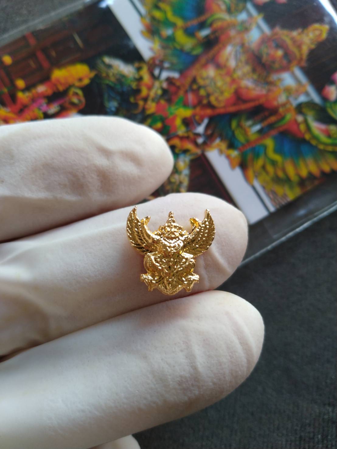 Gloved hand holding a gold mini Garuda amulet pendant with detailed relief and colorful background card