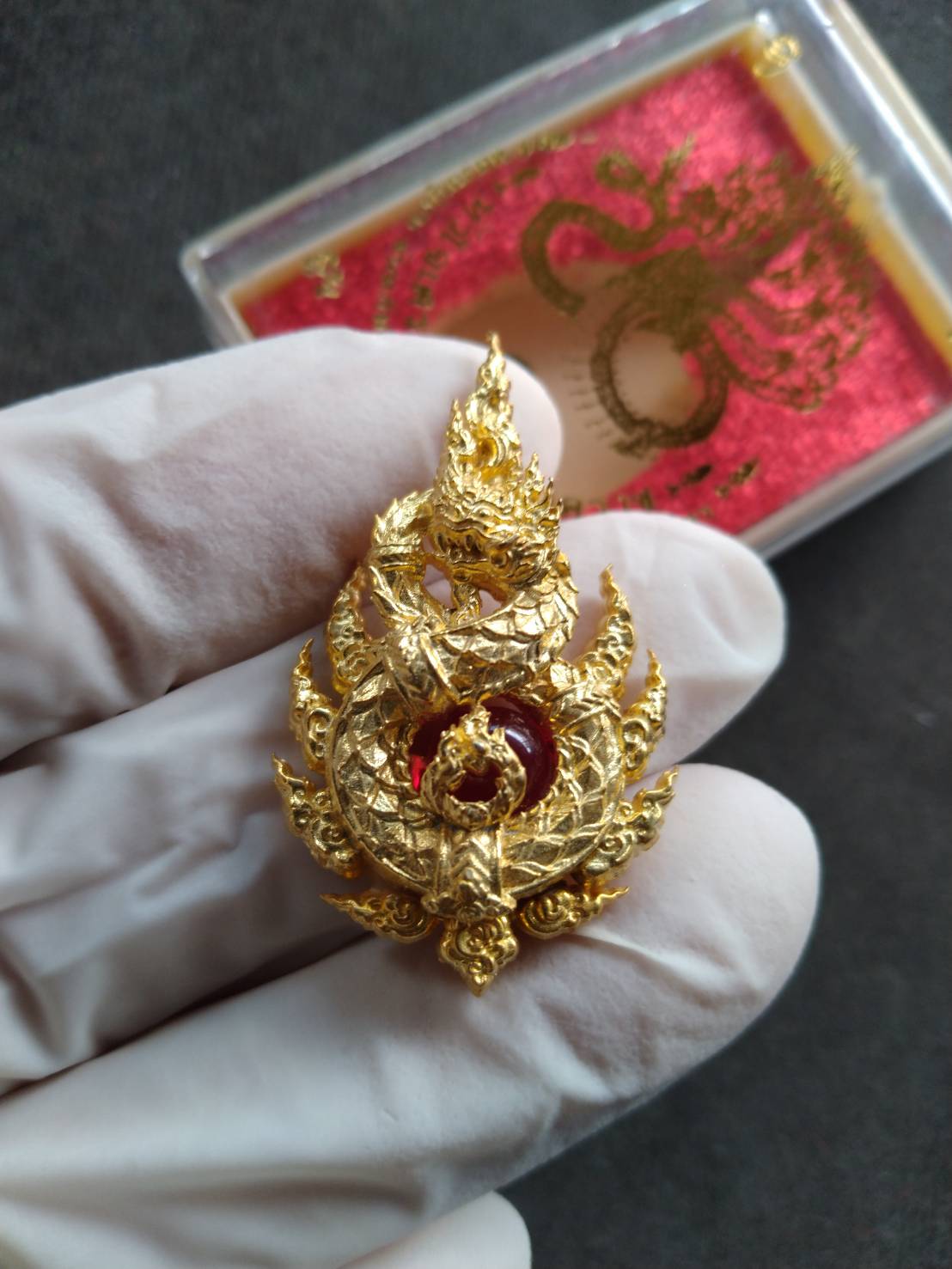 Gloved hand holding gold Naga King Ananta pendant with red gem center and ornate Thai blessing box