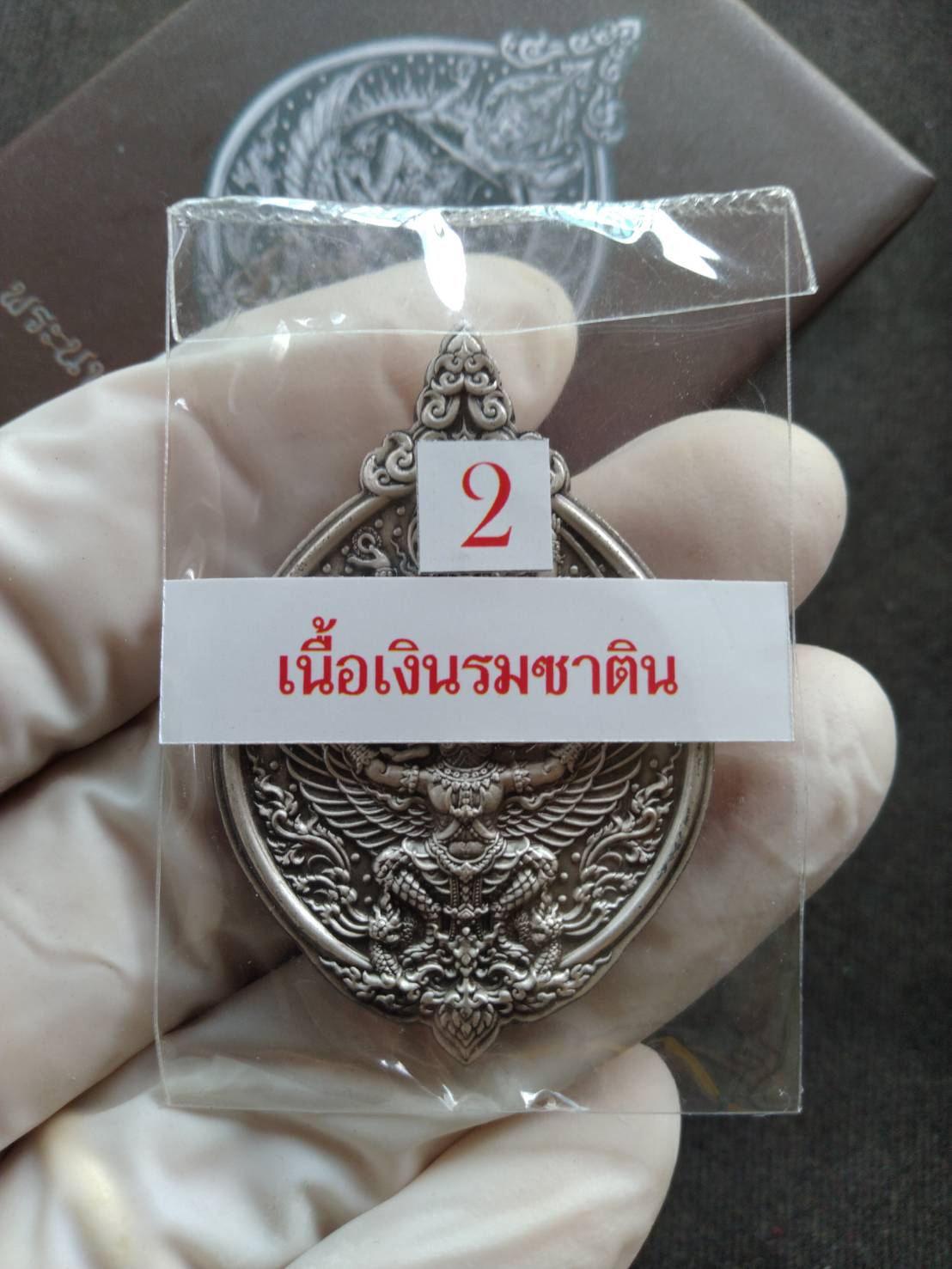 Narayana on Garuda silver amulet packaged in clear pouch held by gloved hand with intricate Thai deity engraving