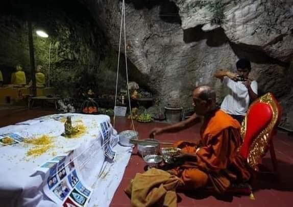 Thai monk blessing Phaya Tao Reuan amulet for wealth on cave altar with offerings and Buddha statues