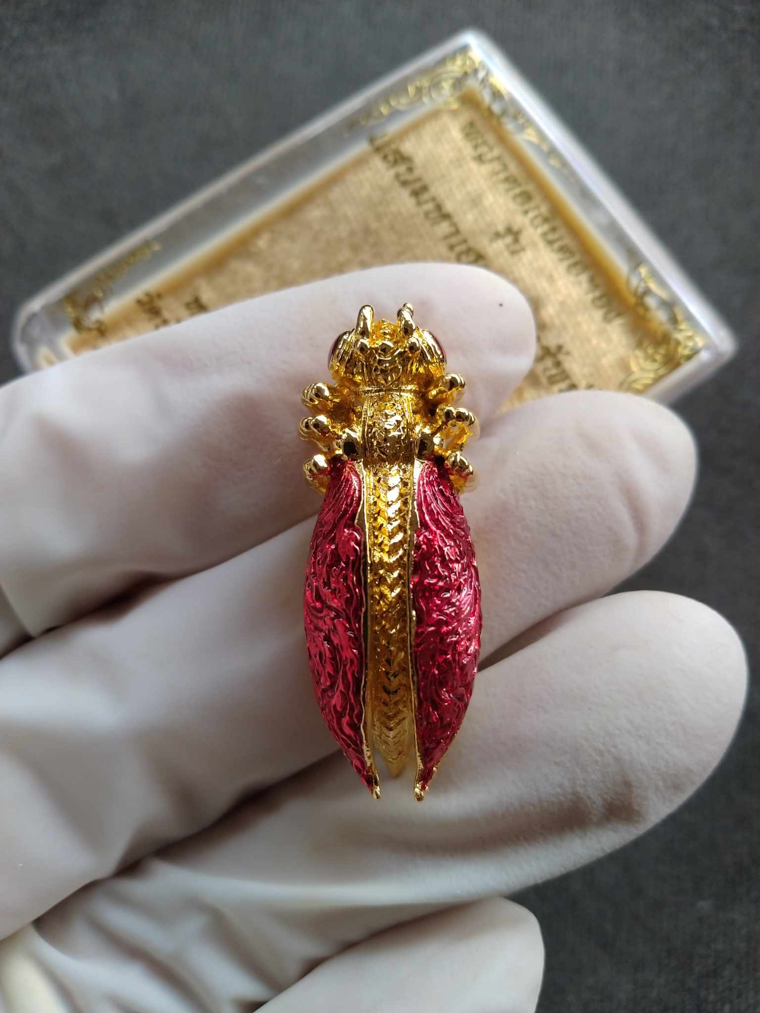 Phaya Tor Ngern Tor Thong Grand Millionaire Amulet  . Gold and red cicada style Thai amulet held in gloved hand by Thai Blessing