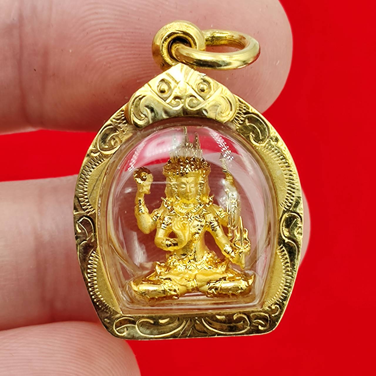 Phra Phrom amulet gold case pendant showing small seated four armed deity in clear domed gold frame on red background