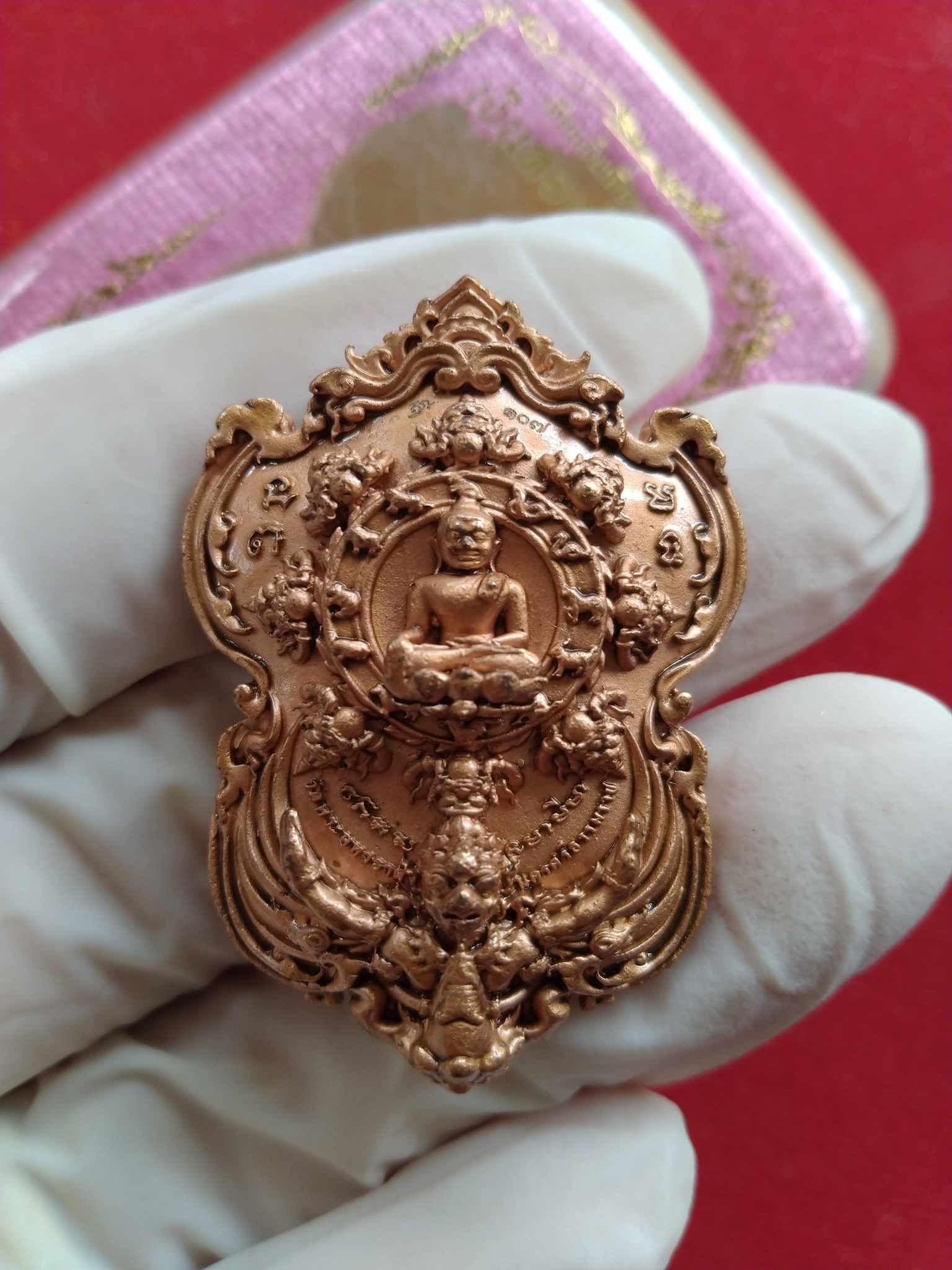 Phra Pidta amulet for wealth copper medallion pendant with ornate Thai Buddha design held in gloved hand