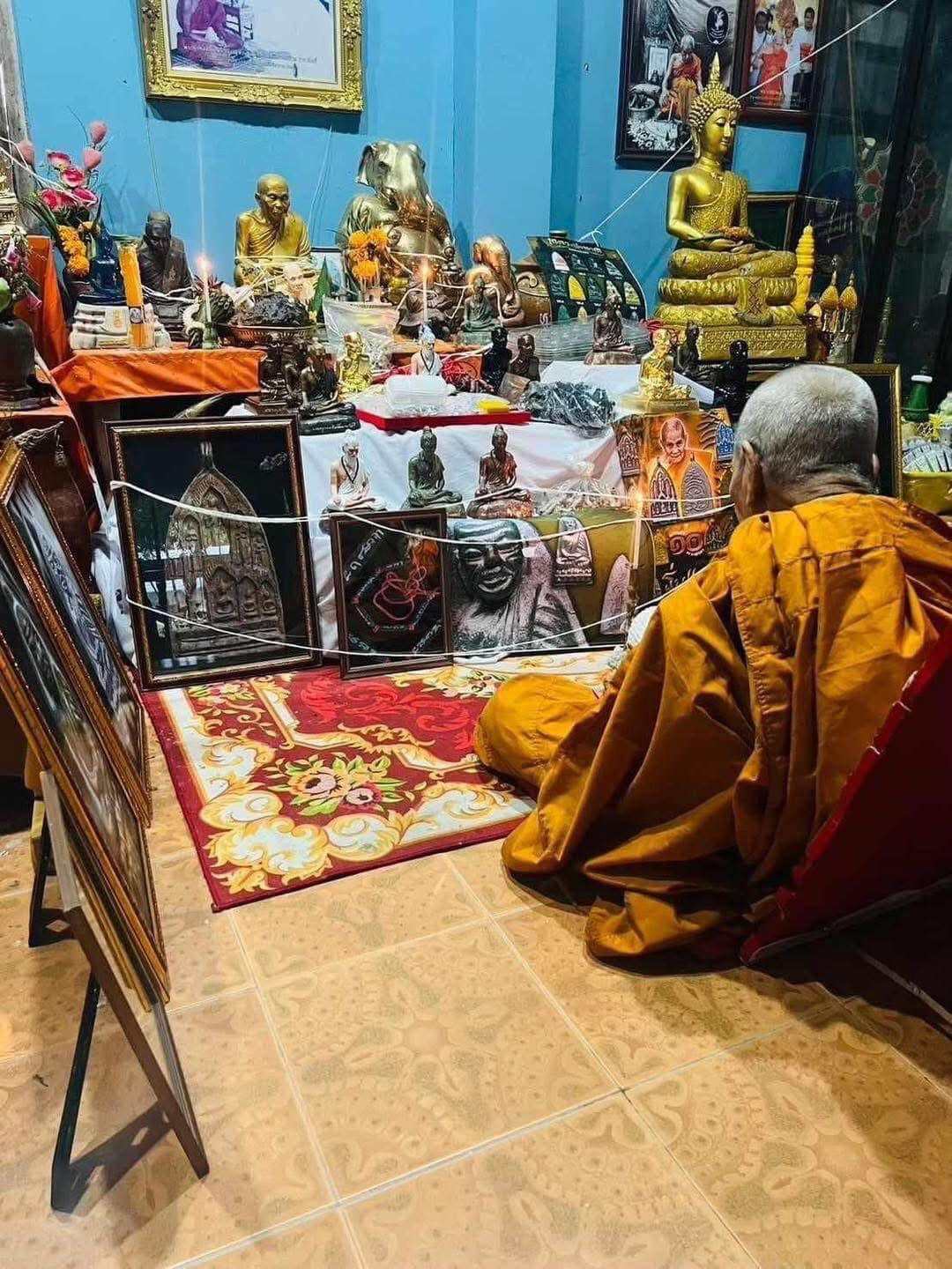 Monk praying at a home shrine with multiple Buddha statues and Phu Lersi Jivaka Komarabhacca statue displayed