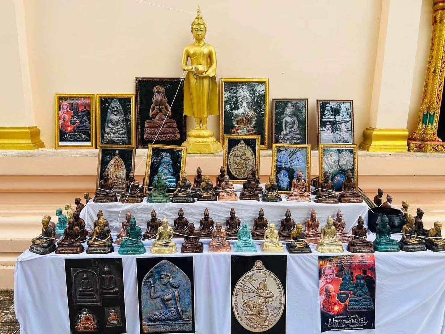Table display of Thai amulets and small Buddha figures with gold standing Buddha in back Phu Lersi Jivaka Komarabhacca statue