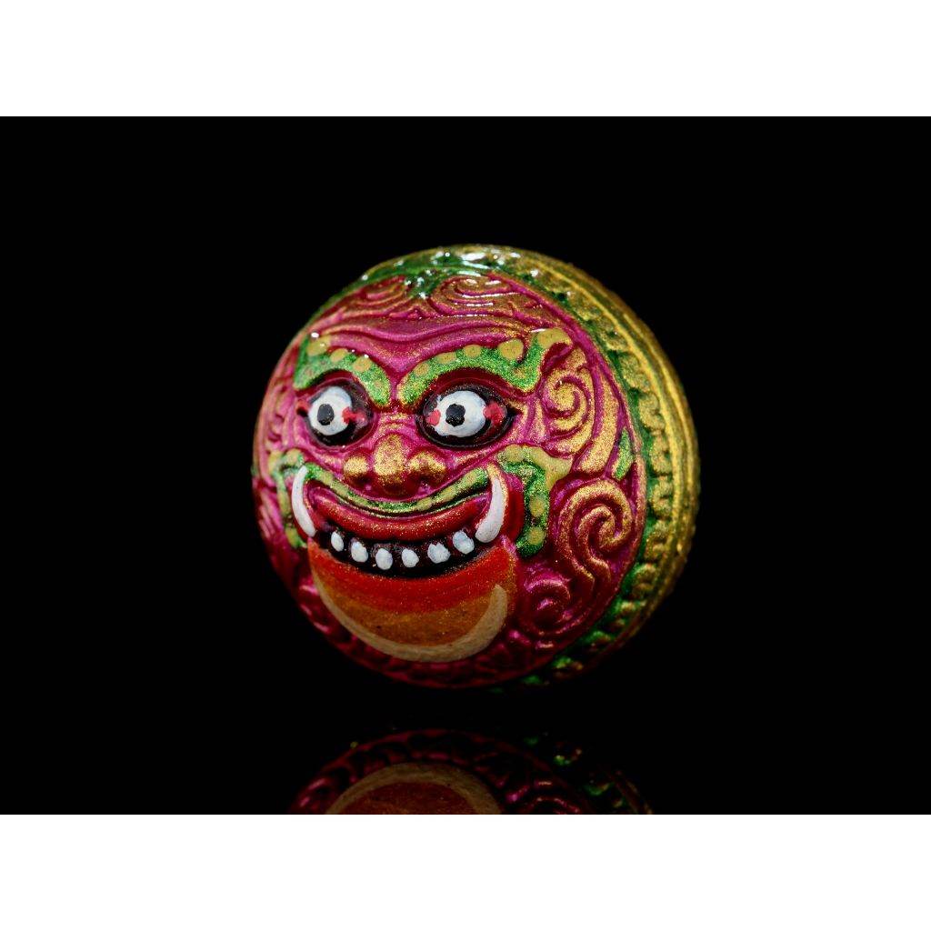 Colorful round Rahu Amulet medallion in pink green and gold featuring a smiling demon face