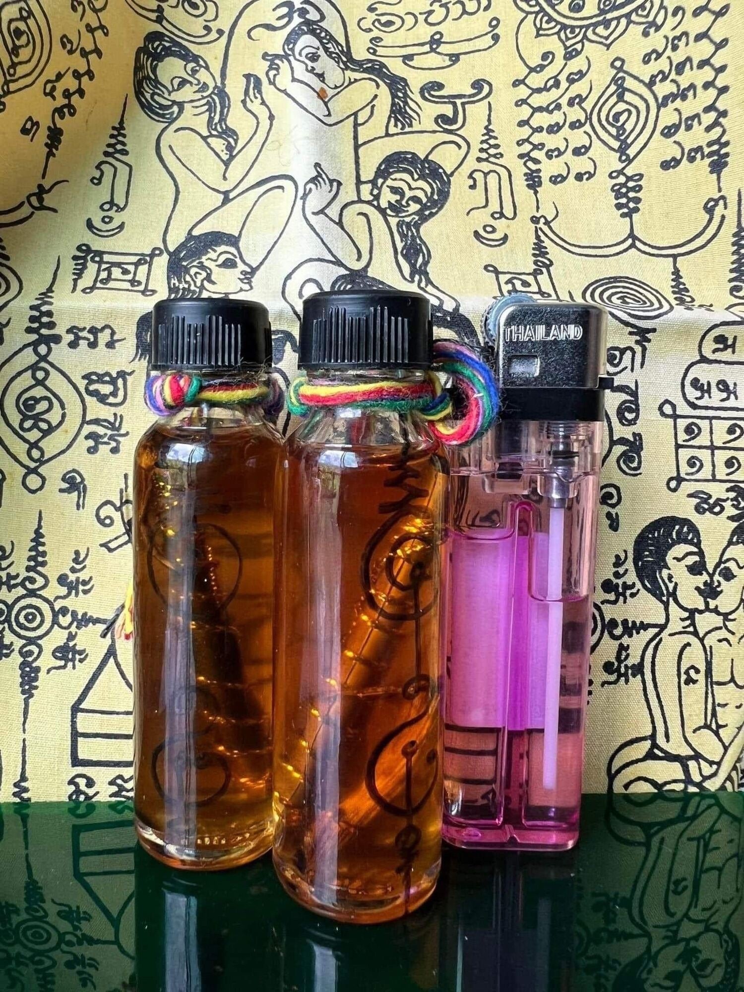 Two small amber bottles with multicolor cord and pink lighter on yantra cloth rare thai attraction oil for relationships