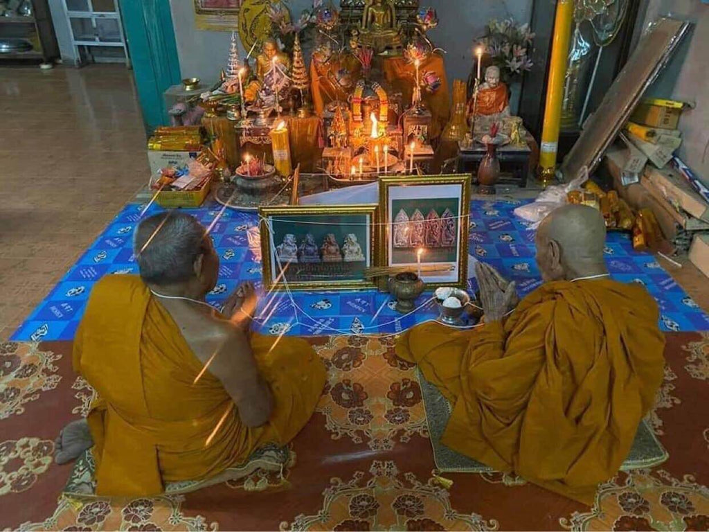 Two Thai monks blessing Ruesi Bai Pai amulet for protection at a home shrine with candles and incense