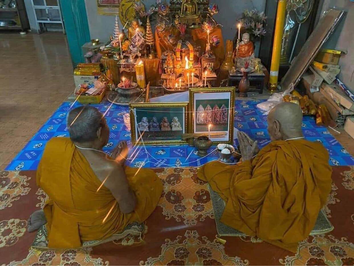 Two Thai monks blessing Ruesi Bai Pai amulet for protection at a home shrine with candles and incense