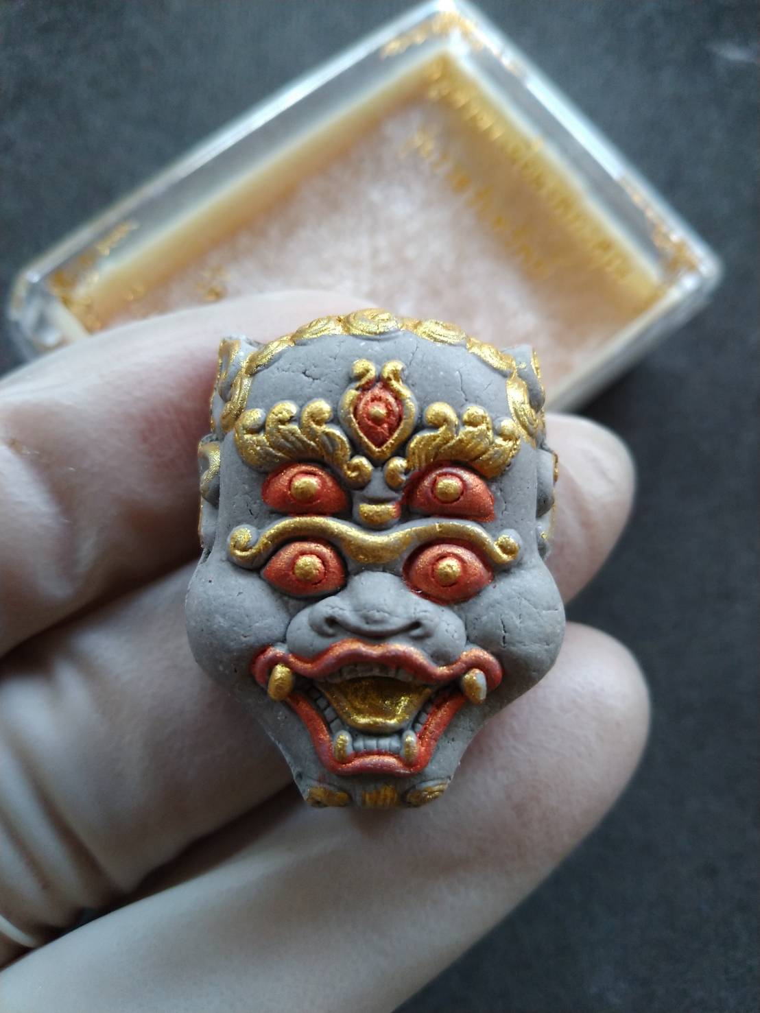 Si Hu Ha Ta prosperity charm Thai carved demon face pendant with gold and red paint held by gloved fingers