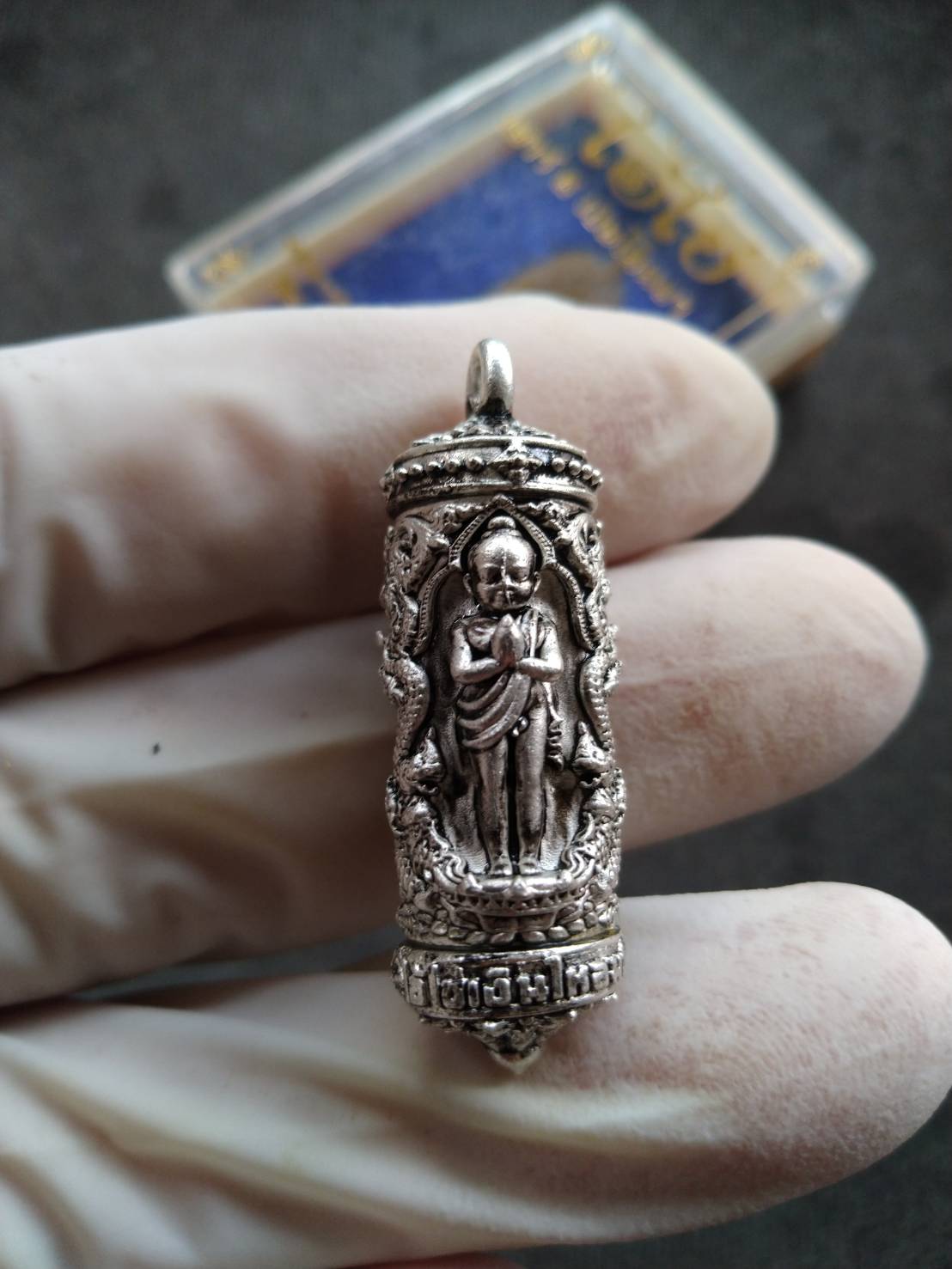 Takrud Ai Khai amulet for wealth silver cylinder pendant with standing monk figure ornate casing held in gloved hand