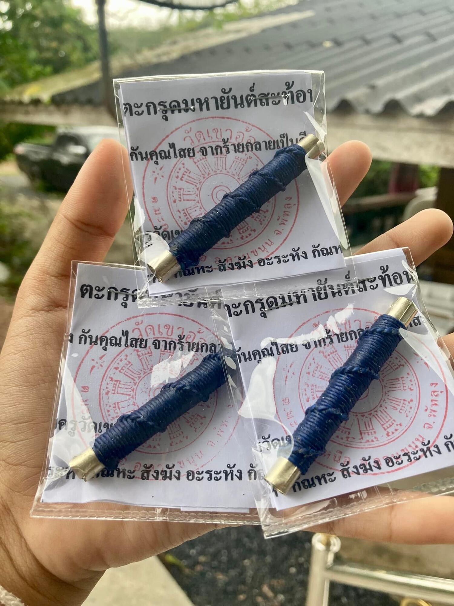 Three blue cord wrapped takruds with metal caps on yantra cards in hand by Thai Blessing Co takrud to reflect negative energy