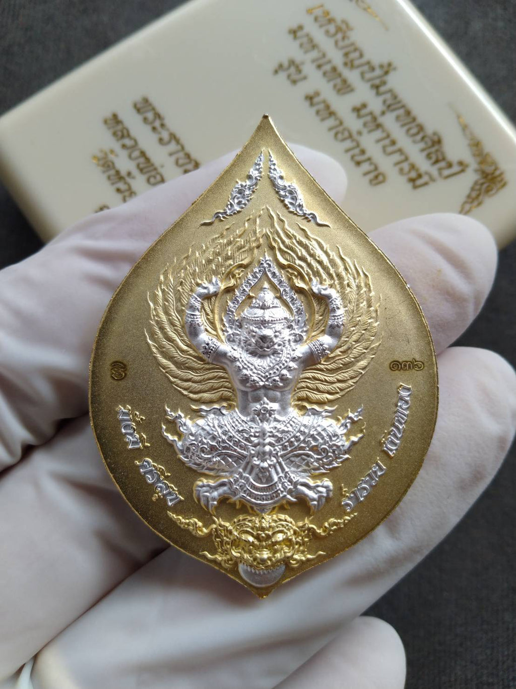 Gold and silver leaf shaped Tao Wessuwan amulet coin with raised guardian deity held in gloved hand