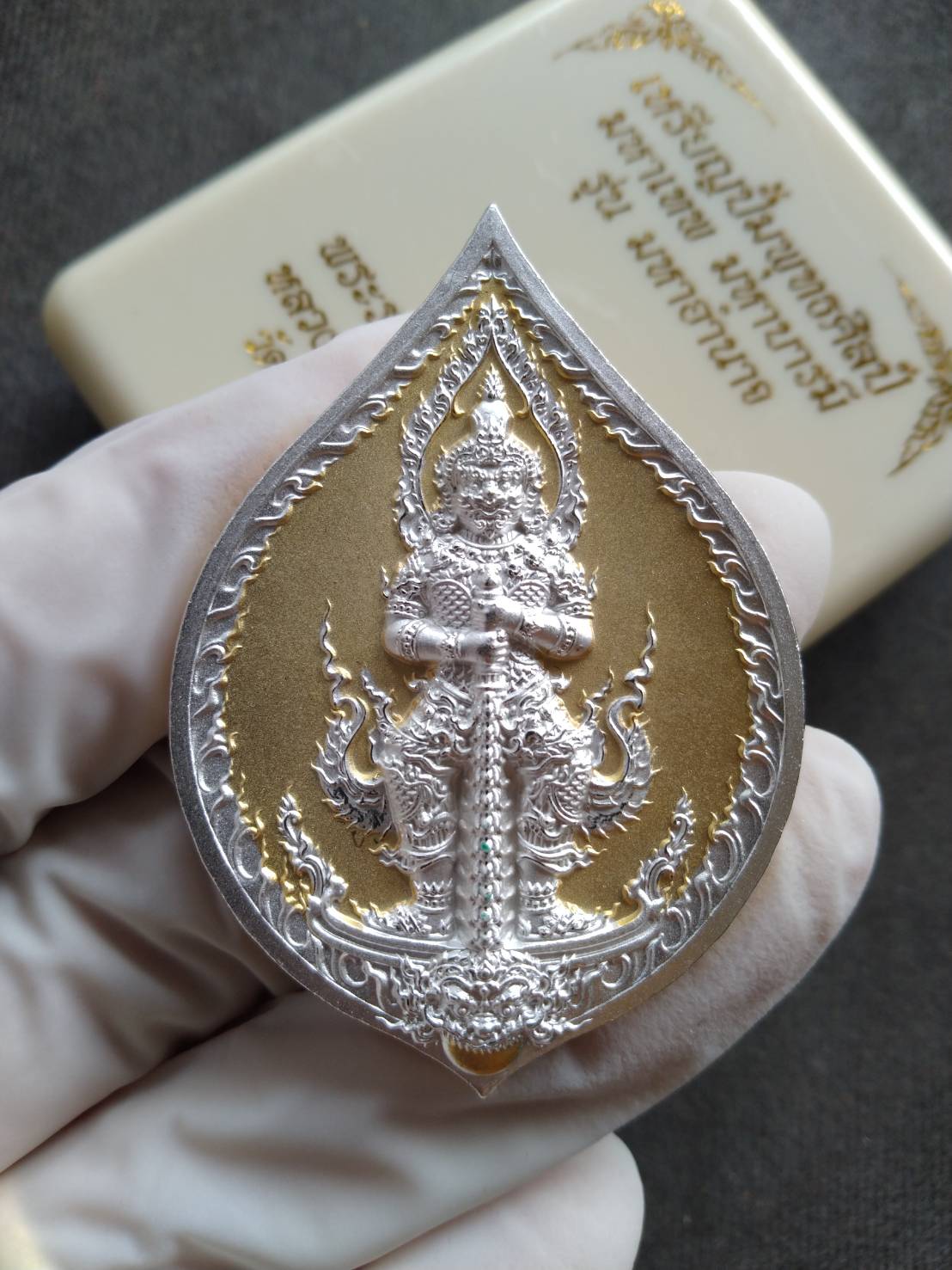 Tao Wessuwan amulet coin silver gold teardrop pendant held in gloved hand, ornate protective guardian deity design