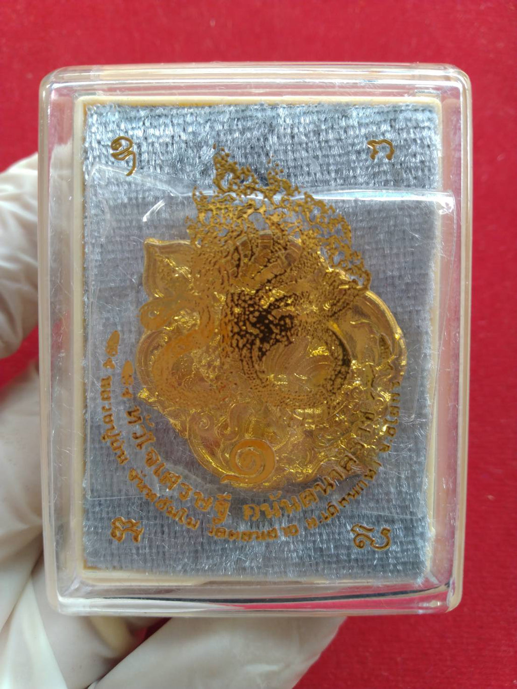 Thai fortune amulet Heart of Wealth gold foil pendant in clear plastic case on gray pad held by gloved hand