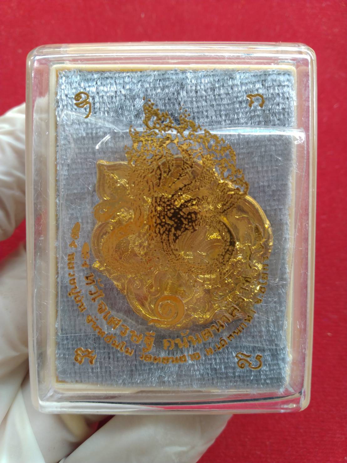 Thai fortune amulet Heart of Wealth gold foil pendant in clear plastic case on gray pad held by gloved hand