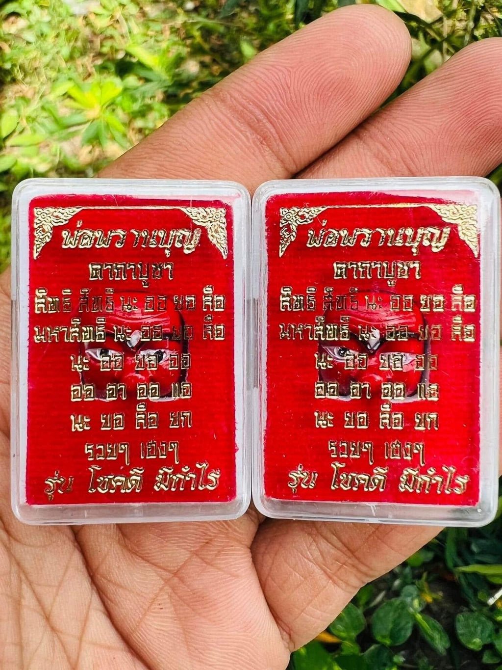 Two red and gold rectangular Thai amulet pendants in clear cases with gold script held in hand Thai prosperity charm for wealth