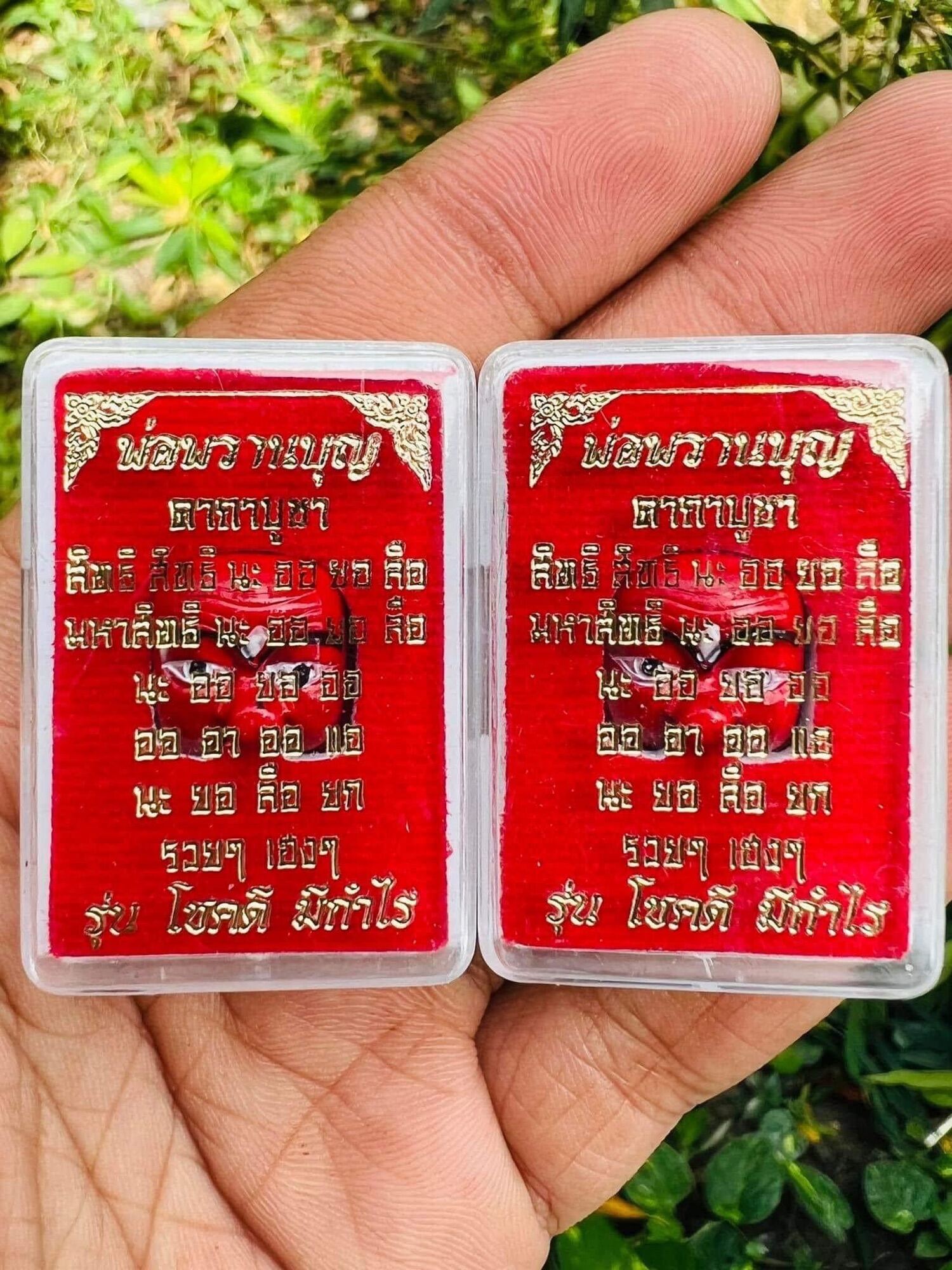 Two red and gold rectangular Thai amulet pendants in clear cases with gold script held in hand Thai prosperity charm for wealth