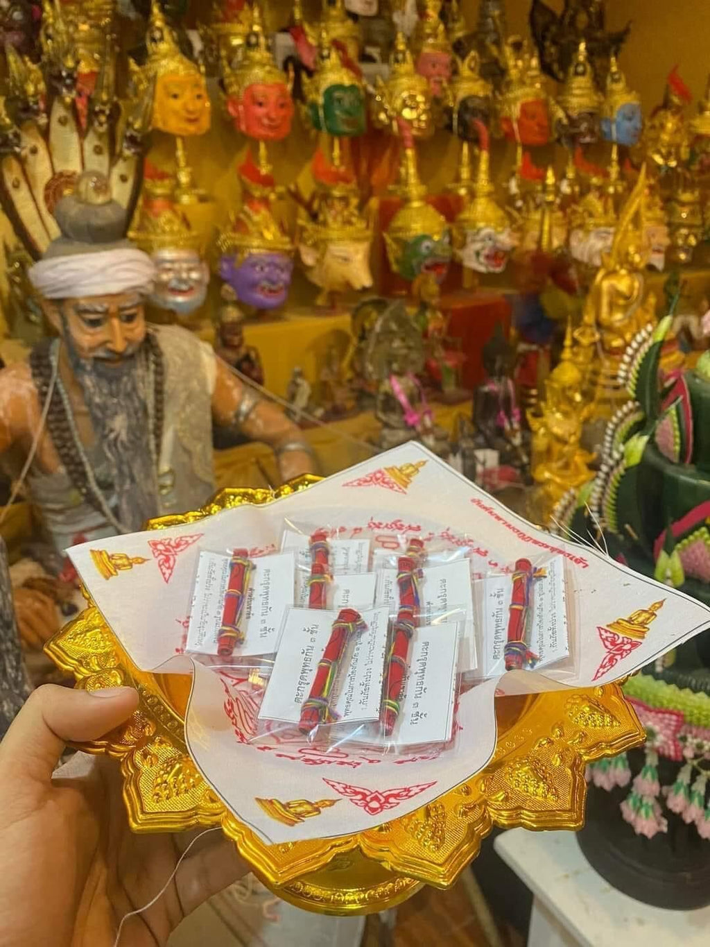 Hand holding golden tray of packaged Triple-layer Phuttha Takrud amulet on cloth with Buddha statues background