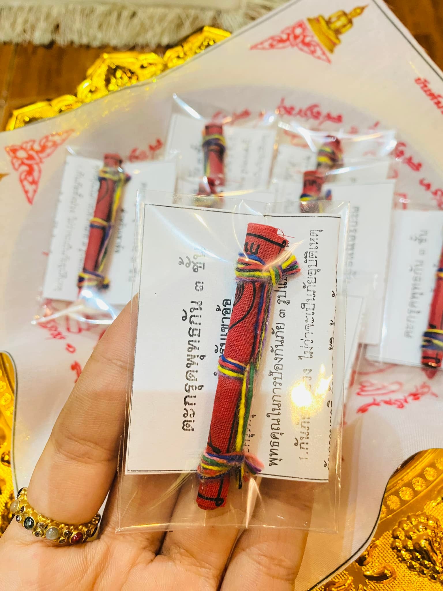 Hand holding Triple-layer Phuttha Takrud amulet wrapped in colorful threads with Thai blessing card and gold tray