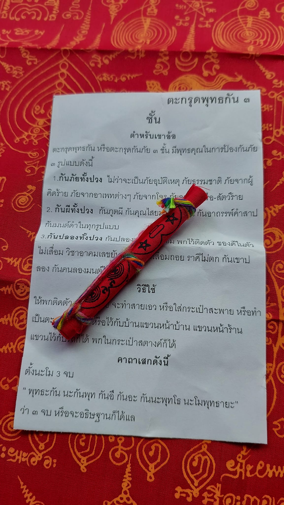 Triple-layer Phuttha Takrud amulet red cloth wrapped scroll on ritual Thai prayer paper over patterned red fabric