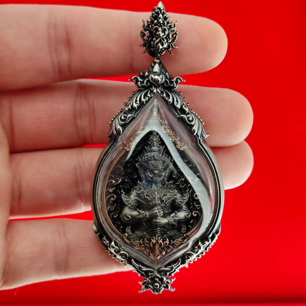 Hand holding ornate silver teardrop pendant with carved deity inside, Lord Vesuvananno amulet for wealth and protection.