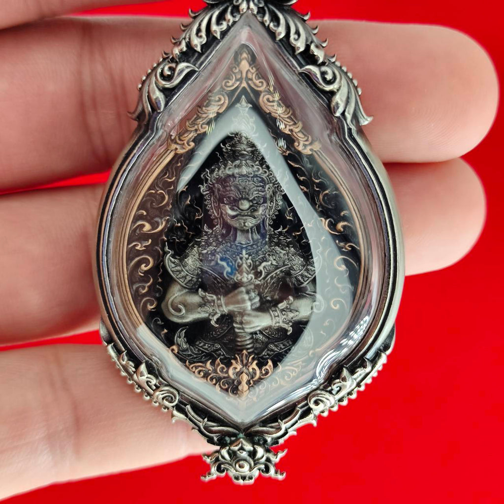 Handheld ornate silver pendant with deity in glass case on red background Lord Vesuvananno amulet for wealth and protection.