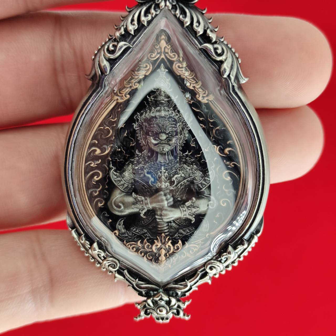 Handheld ornate silver pendant with deity in glass case on red background Lord Vesuvananno amulet for wealth and protection.