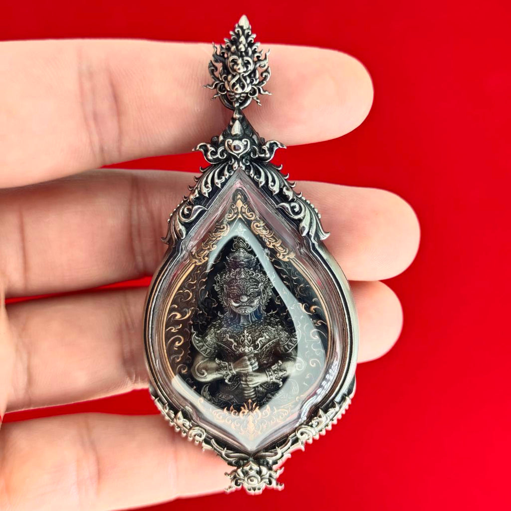 Hand holding ornate silver teardrop with deity figure amulet on red backdrop Lord Vesuvananno amulet for wealth and protection.