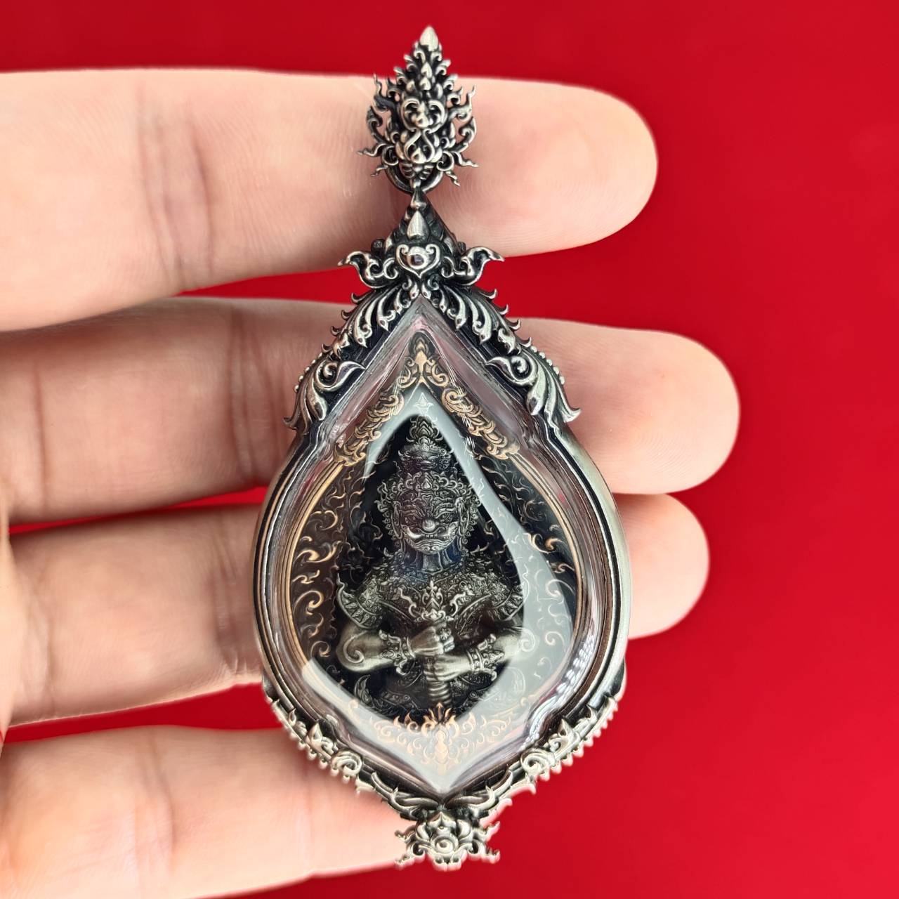 Hand holding ornate silver teardrop with deity figure amulet on red backdrop Lord Vesuvananno amulet for wealth and protection.