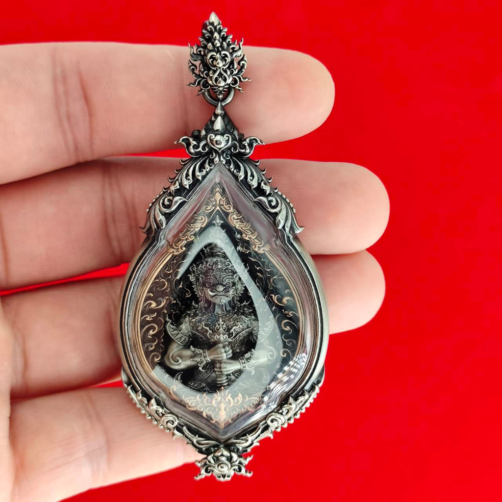 Hand holding ornate teardrop silver pendant with deity inside on red background, Lord Vesuvananno amulet for wealth and protection.