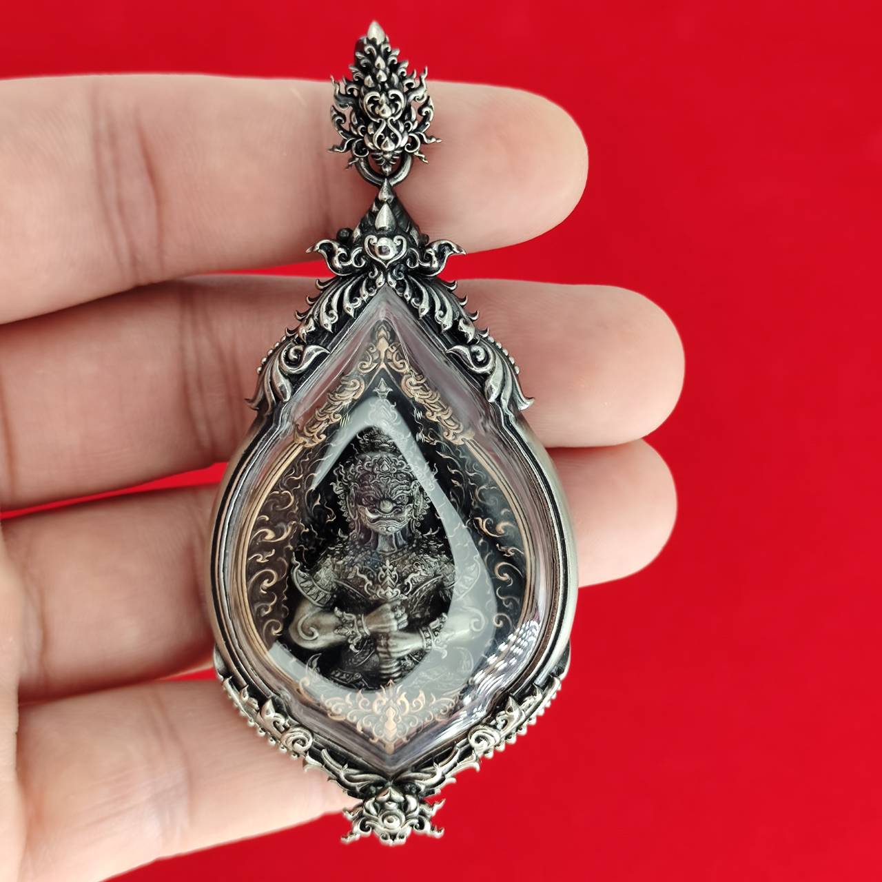Hand holding ornate teardrop silver pendant with deity inside on red background, Lord Vesuvananno amulet for wealth and protection.
