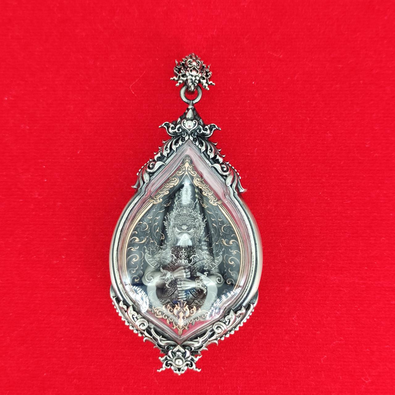Thai Blessing Lord Vesuvananno amulet for wealth and protection. Ornate silver teardrop amulet in display case on red background