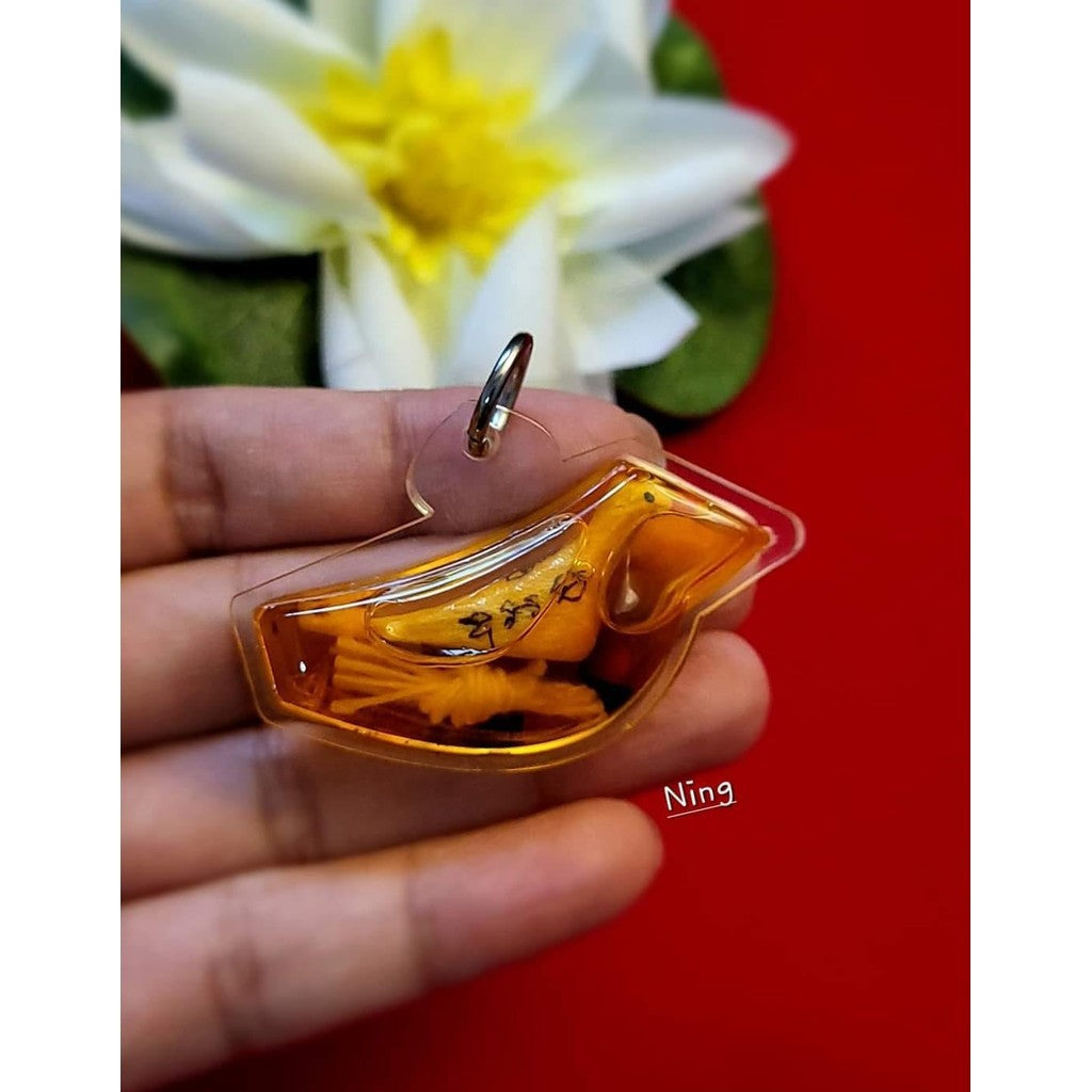 Hand holding a Thai blessed lucky charm keychain with gold and amber colors in front of a white yellow flower