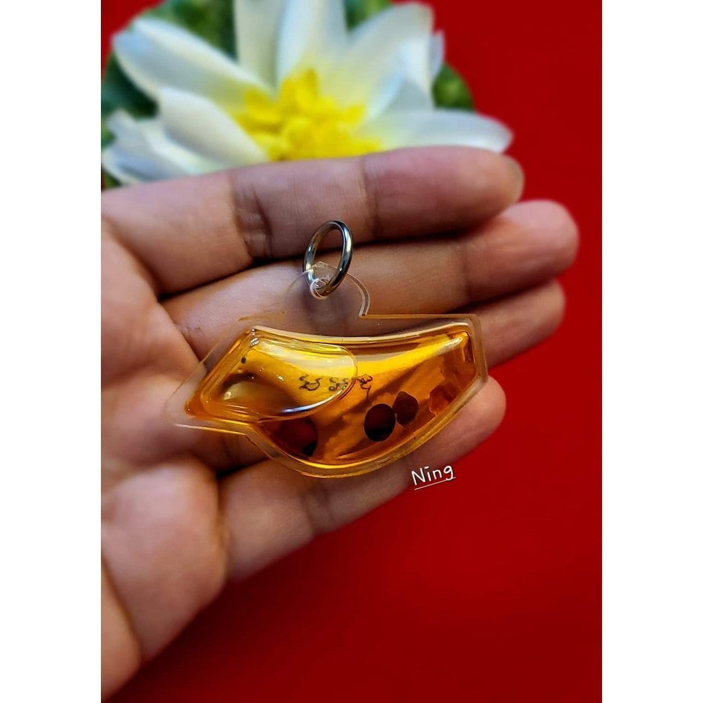Hand holding a Thai blessed lucky charm in amber color with black dots and script inside a clear casing