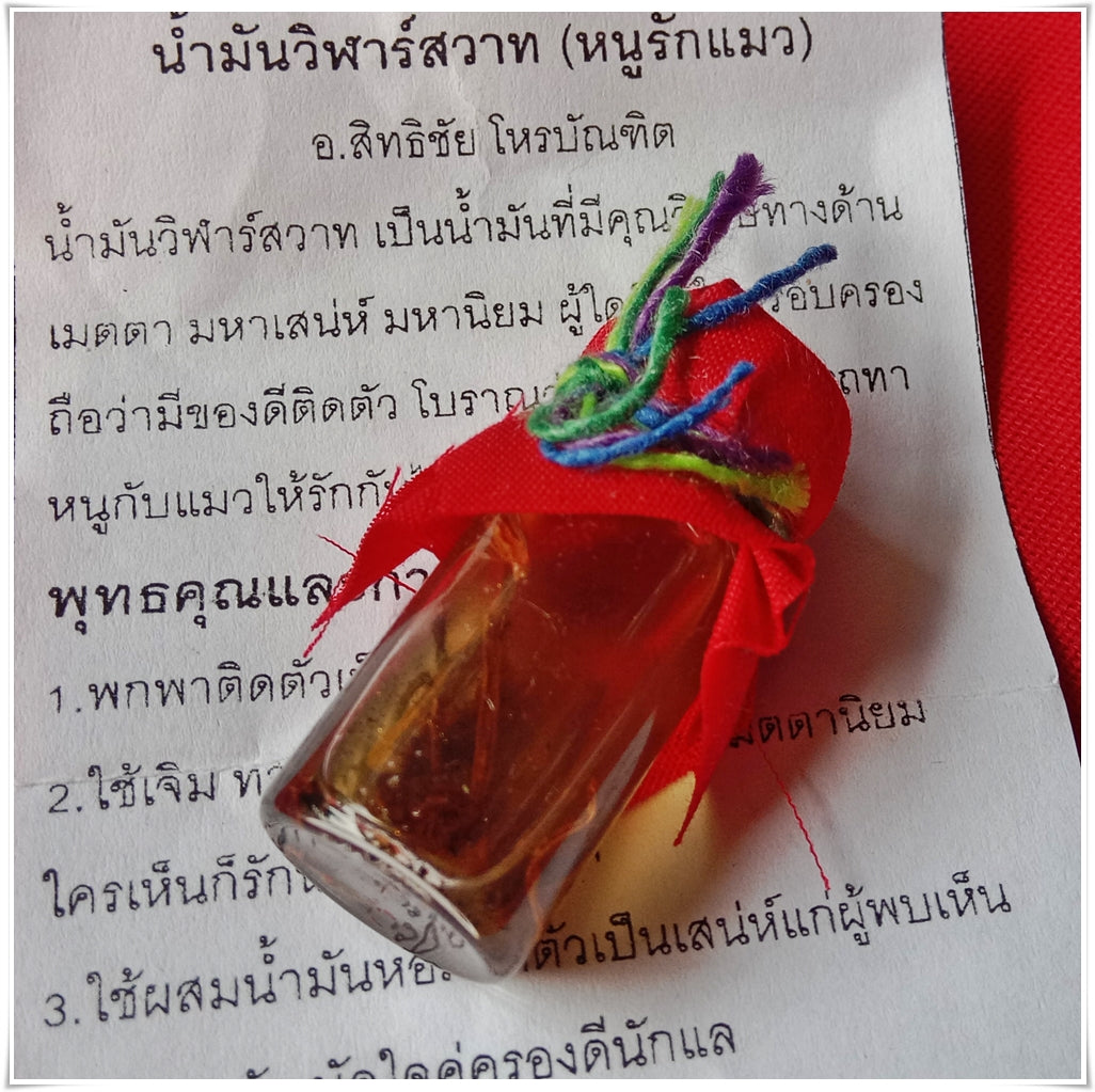 Small glass vial of Thai Buddhist charm oil for loving kindness wrapped in red cloth with multicolor yarn atop Thai script paper