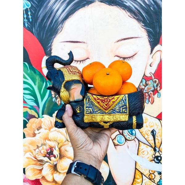 Hand holding a Thai elephant resin tray filled with fresh oranges against colorful mural background