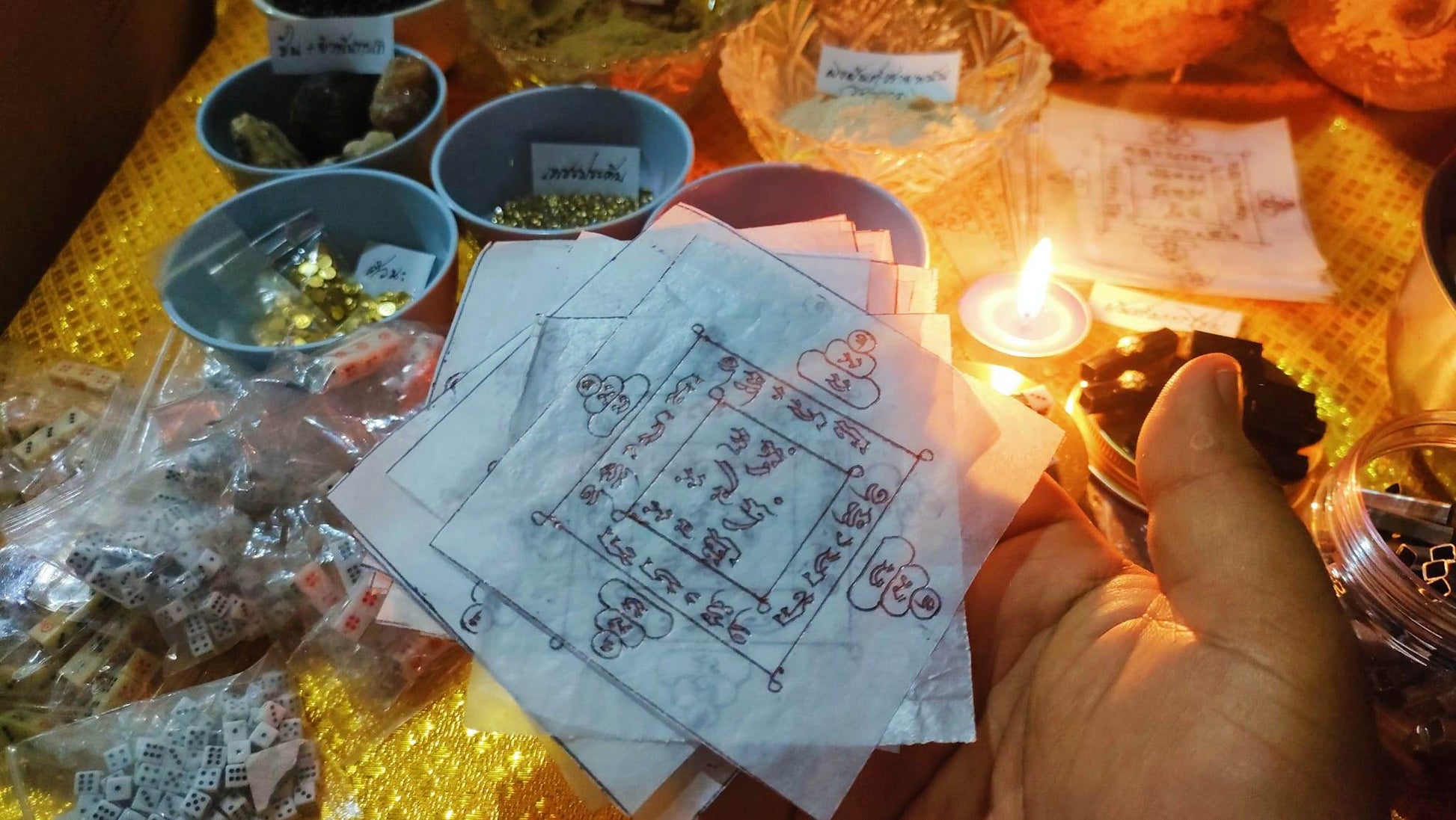 Hand holding translucent paper amulets with handwritten symbols of Thai gambler's takrud amulet surrounded by ritual items