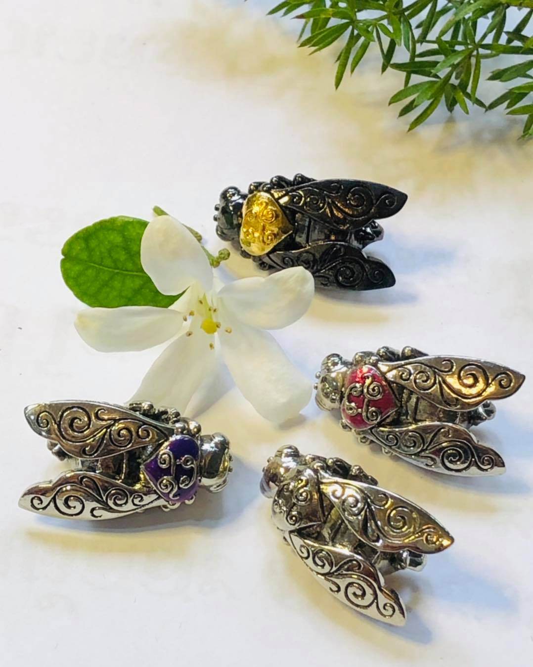 Thai golden bee amulet for wealth in silver-tone with intricate designs and colorful accents displayed with a white flower and green leaves