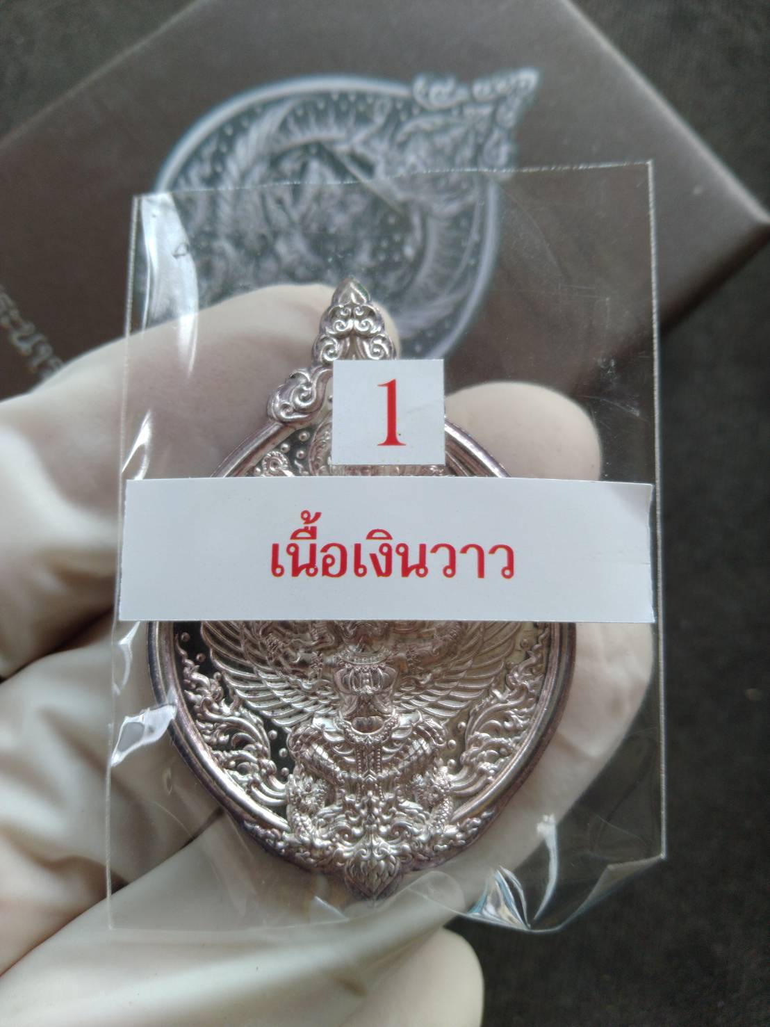 Narai on Garuda silver amulet in clear plastic packaging held by gloved hand showing intricate embossed Thai design